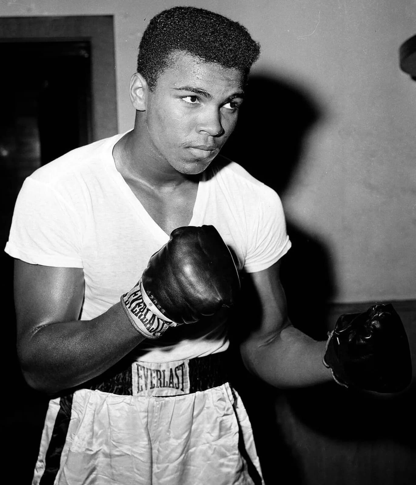 A young Cassius Clay in a tight white T-shirt trains at the City Parks Gym in New York in 1962, fists raised, watched by onlookers.