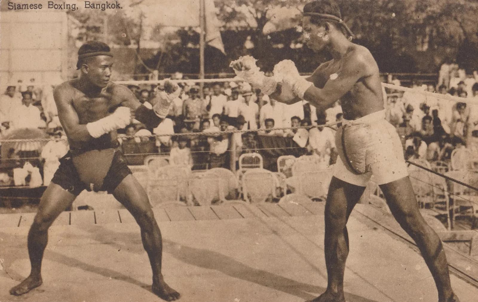 Two Siamese boxers with rope-bound fists face each other in an outdoor ring in Bangkok in the 1920s, a crowd watching.