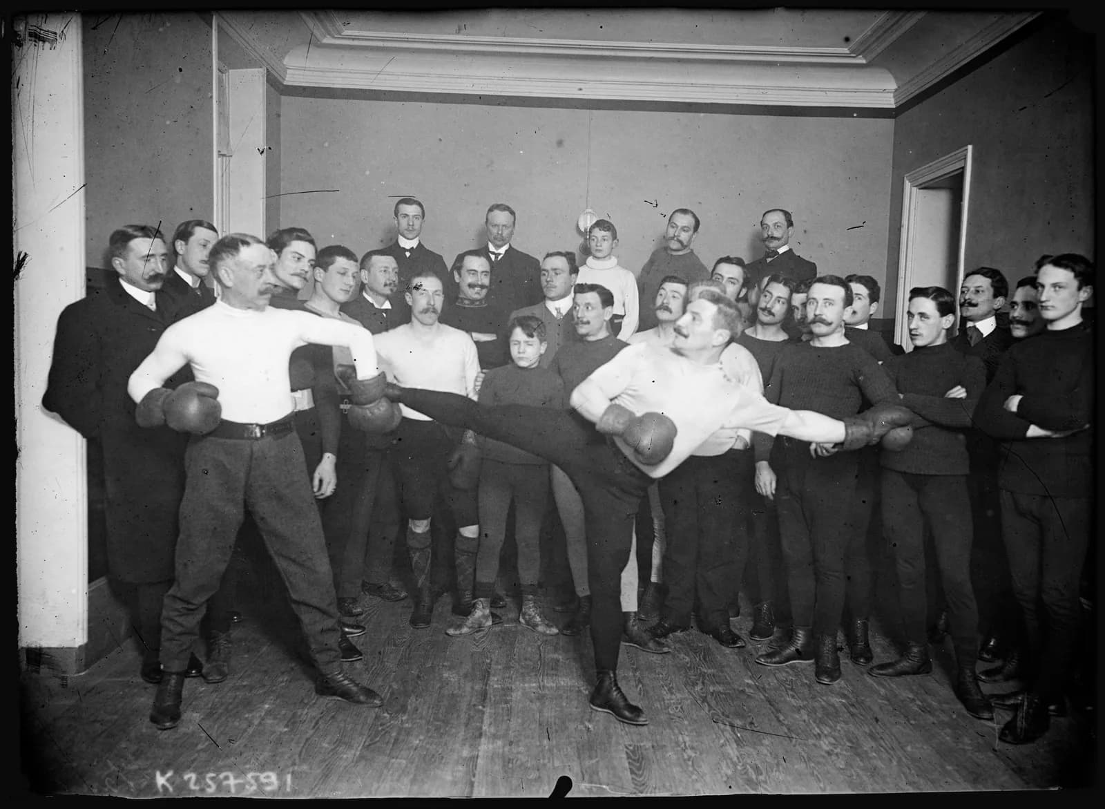 Photograph from 1906 of the savate teacher Castérès demonstrating a high side kick against a partner in a gymnasium.