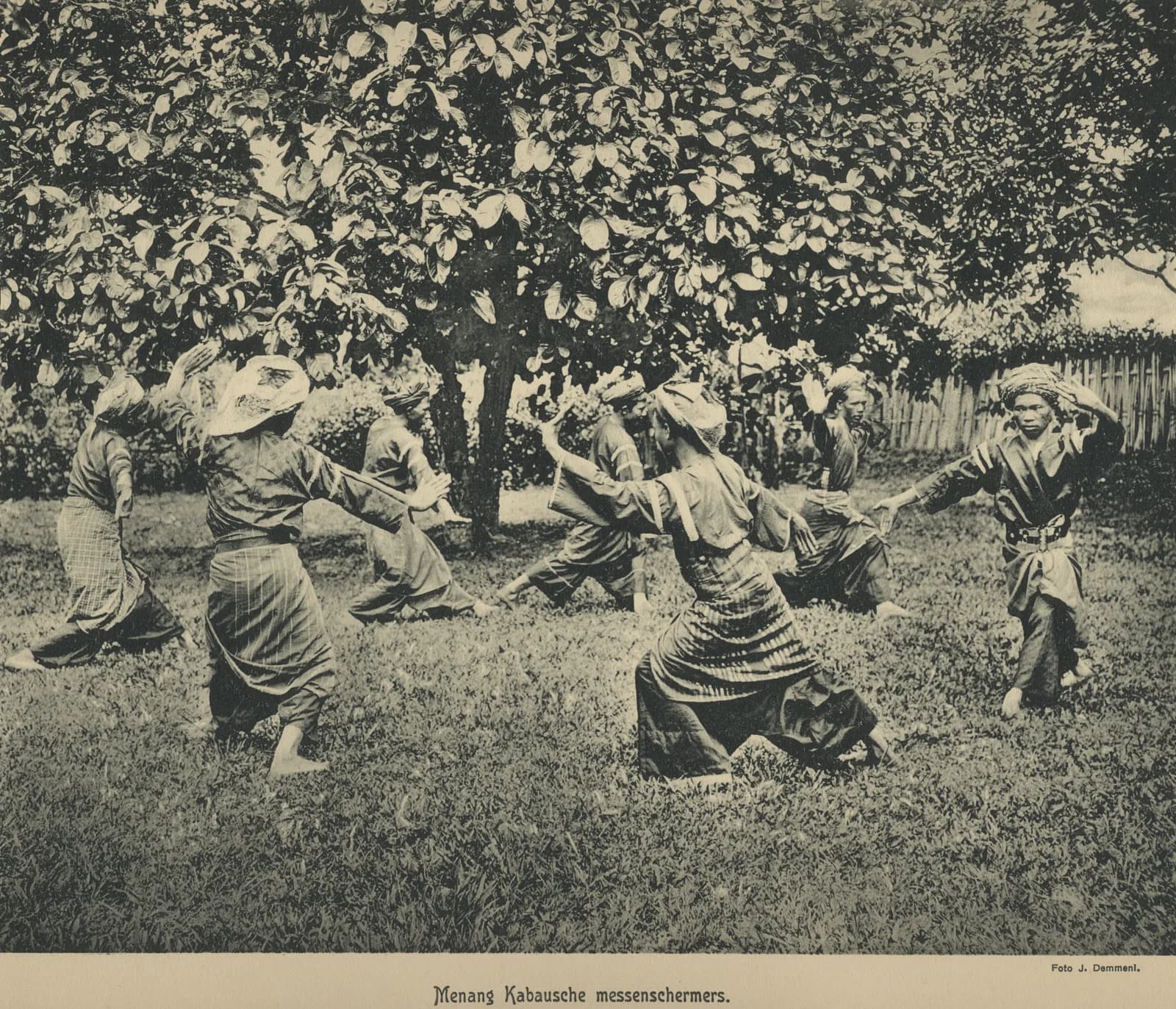 Photograph from around 1915 of two Minangkabau men in traditional dress practising silat in a low stance, watched by villagers on Sumatra's west coast.